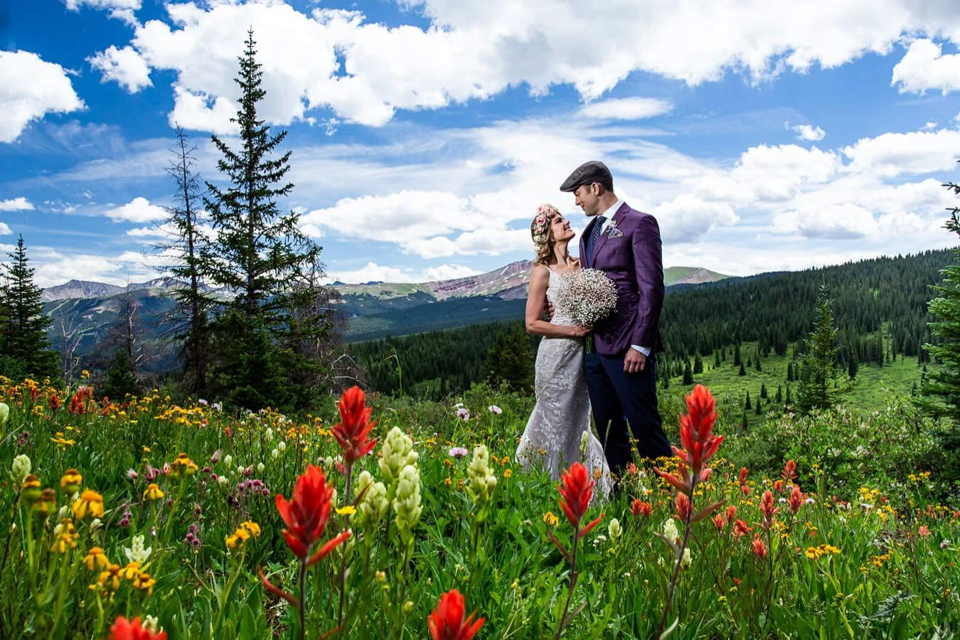 A bride wearing a flower crown looks up at the groom wearing a purple suit, surrounded by red, yellow and white wildflowers in Vail, Colorado with mountains in the background.
