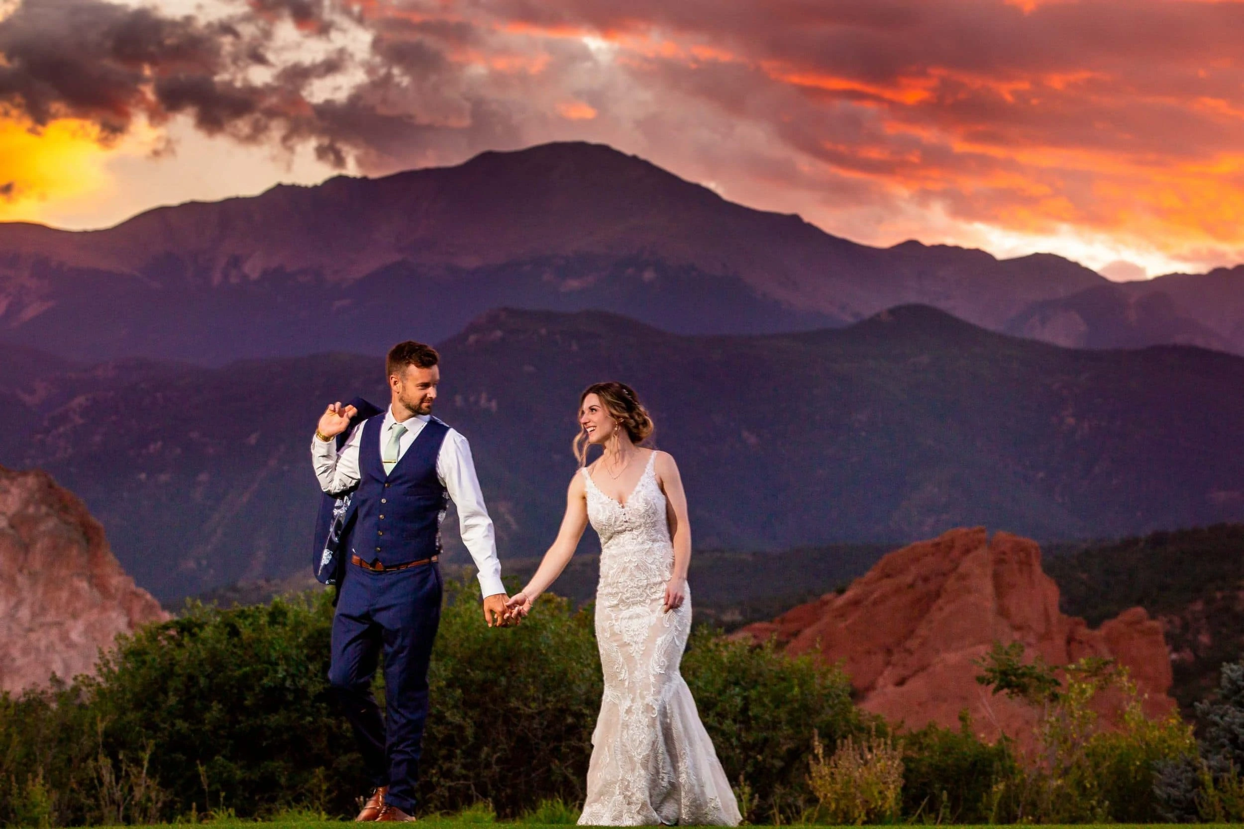 A bride and groom walk hand in hand at sunset with Garden of the Gods in the background.