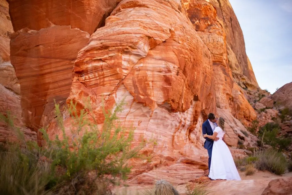 Valley of fire elopement photographer Lucy Schultz shows off her colorful and vibrant style as a groom in a navy suit kisses his wife in a white dress in front of a huge red rock.
