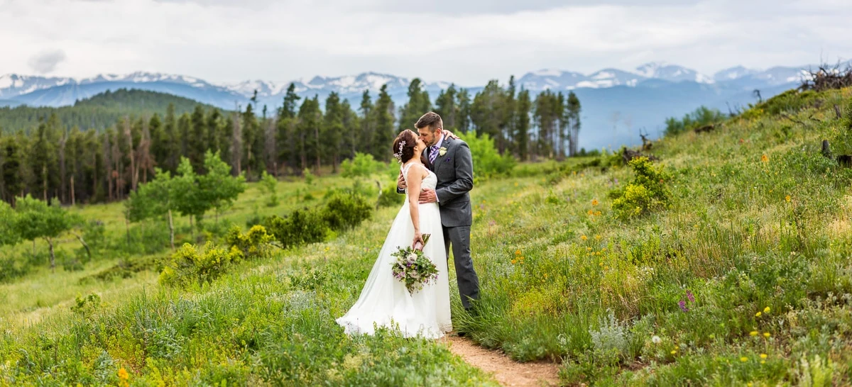 An elopement couple kisses in a field of colorful flowers in Nederland, . Snow capped mountains are visible above the pine trees.