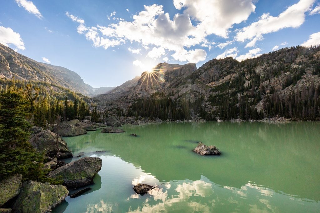 A colorful landscape photo of the greenish waters of Lake Haiyaha in Rocky Mountain National Park. The sunburst on the horizon touches Hallett's peak.