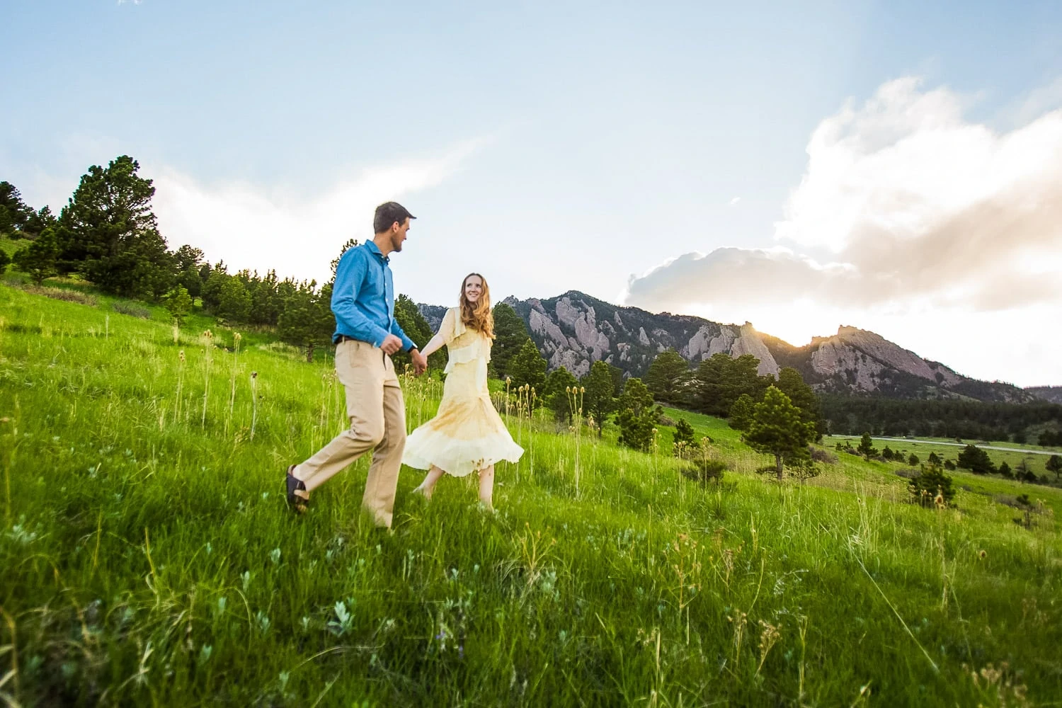 A bride in a vintage dress walks with her husband down a green hill in front of the Boulder Flatirons.