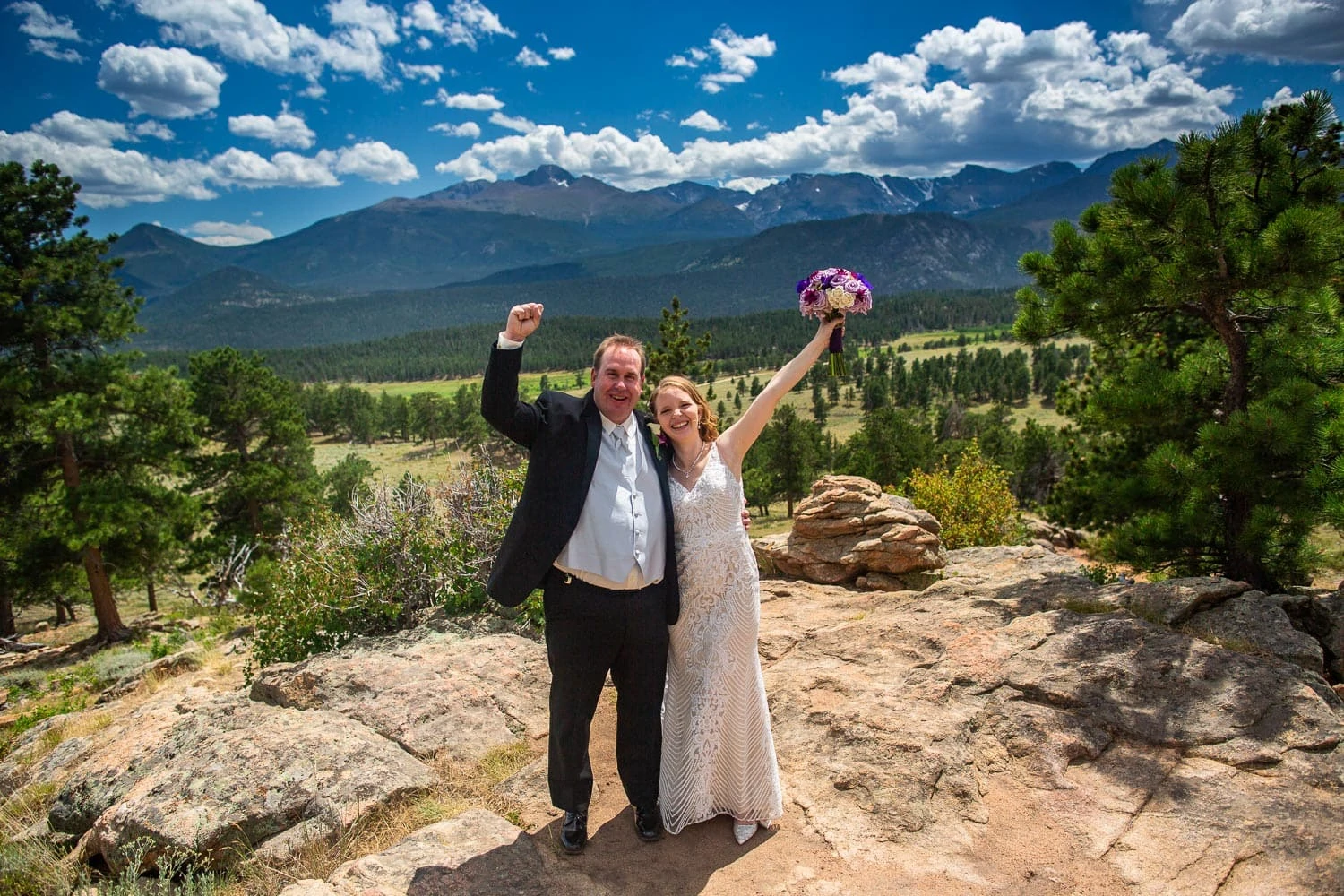 A newlywed couple lift their arms to the sky in triumph after their Rocky Mountain National park elopement at 3m curve in summer.