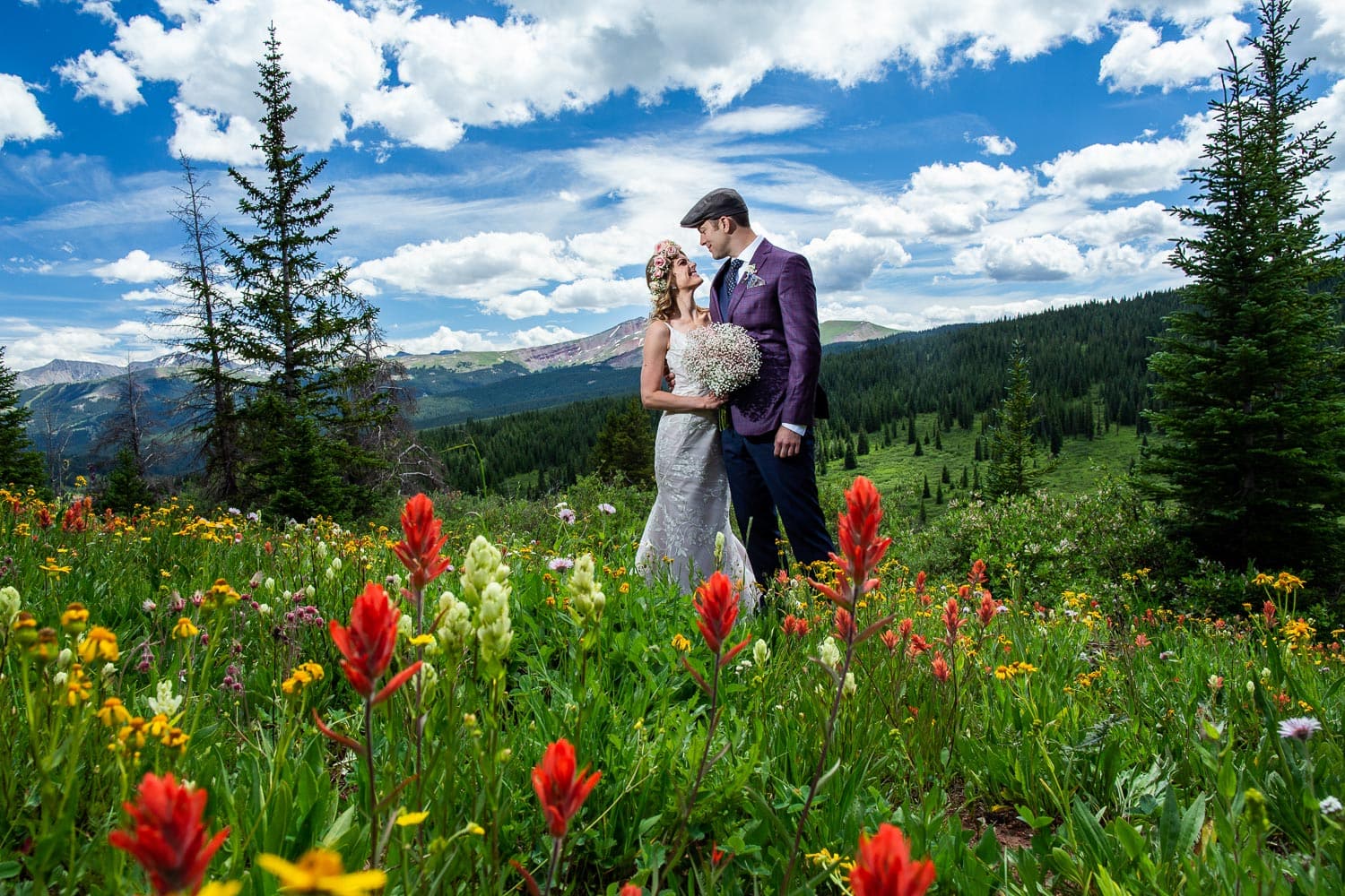 A boho bride and a groom in a purple jacket on the side of a mountain covered in wildflowers.