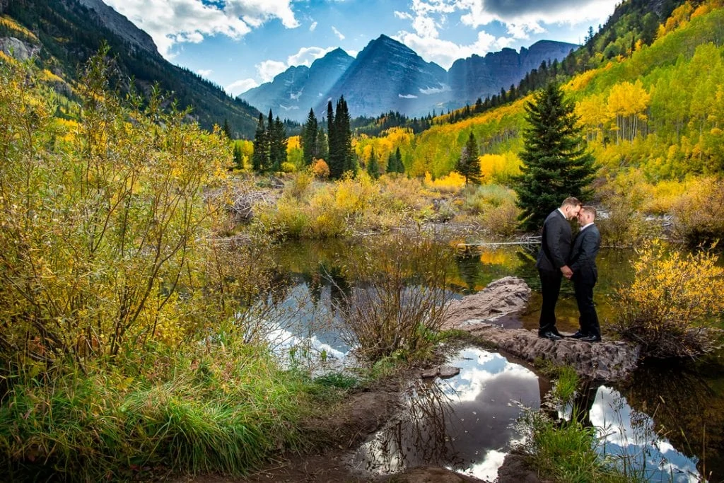 A gay elopement couple stands on a small rock in a lake in front of the Maroon Bells peaks in Aspen, Colorado. Light is streaming down from the clouds over them.