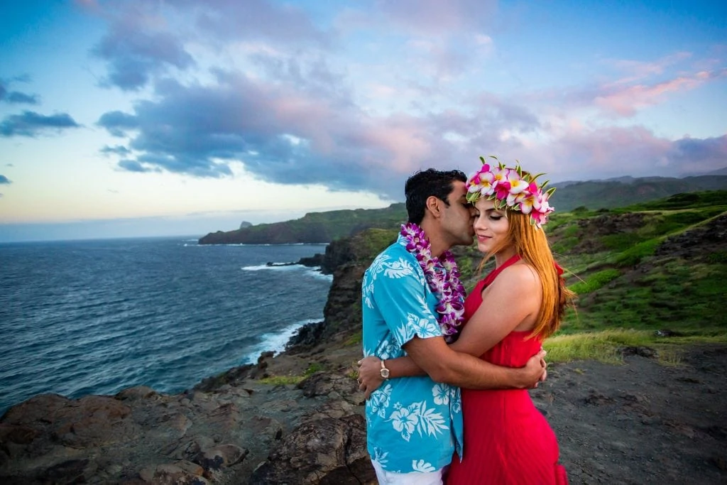 A Maui elopement couple smooches on the green coastline. He is wearing a blue Hawaiian shirt and purple lei and she is wearing a bright red dress and pink flowers in her hair.