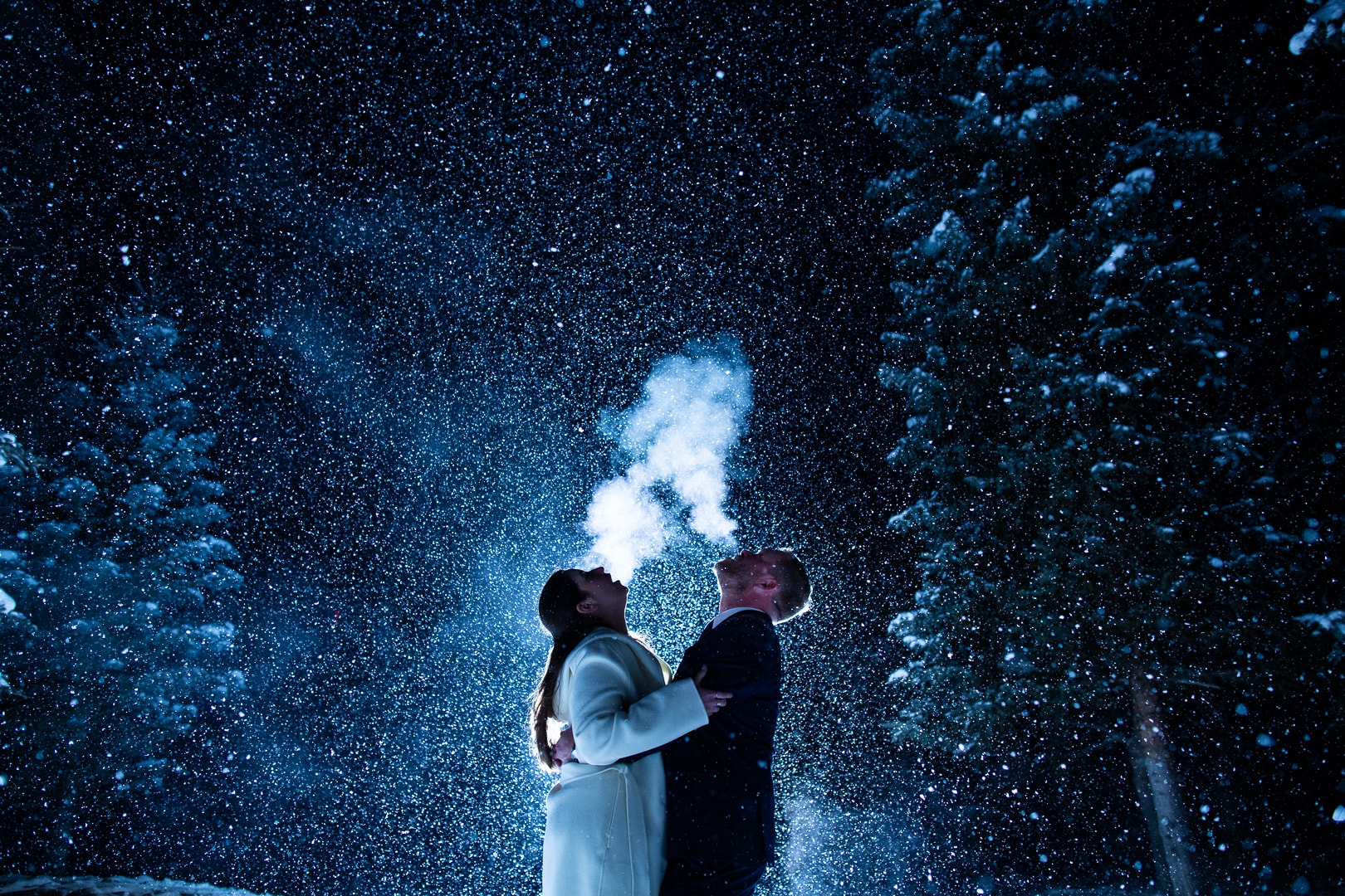 A bride and groom see their breath as snow falls around them after dark.