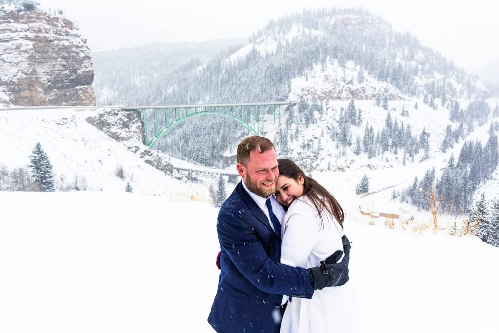 A wedding couple wearing coats and gloves embrace in front of a snowy landscape during their winter elopement.