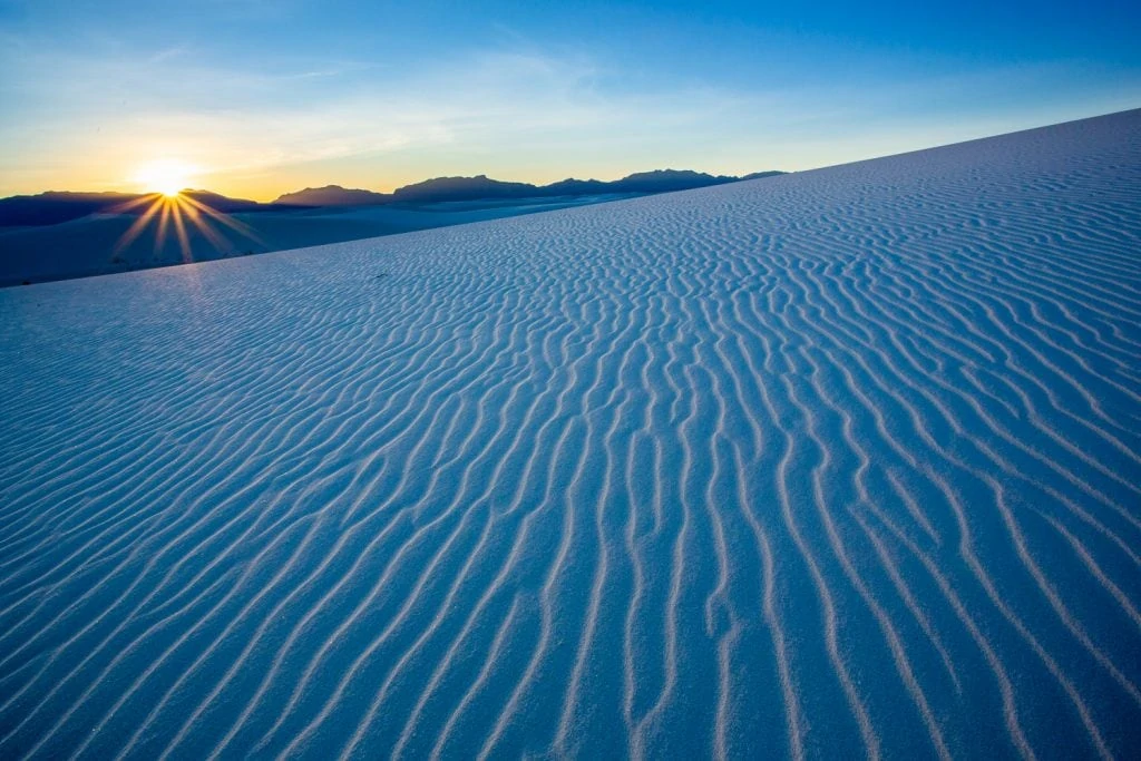 A view of rippled white sand dune as the sun sets over the mountains at White Sands National park.