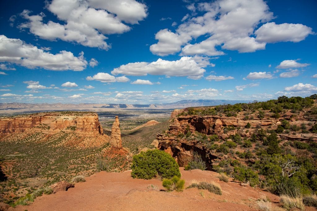 The red canyon walls of Colorado National Monument in a dramatic view of the canyon, with its gulches, spires, and rock formations dotted with desert vegetation.