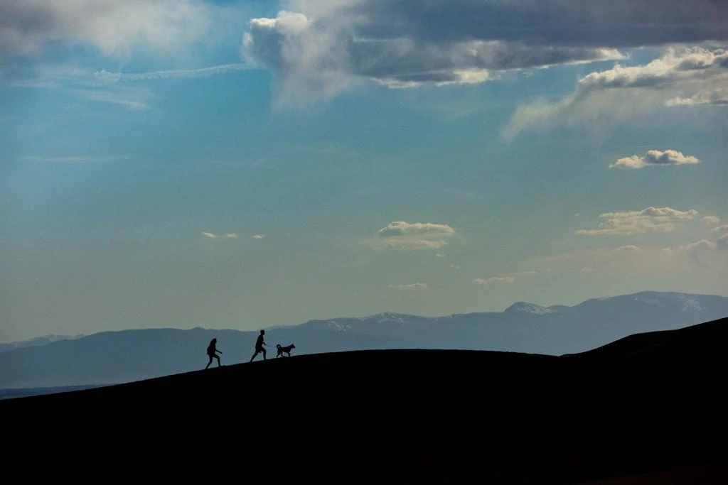 A silhouette of a couple and their dog on the spine of a dune with mountains in the background. Taken at Great Sand Dunes National Park where dogs are allowed on leash.