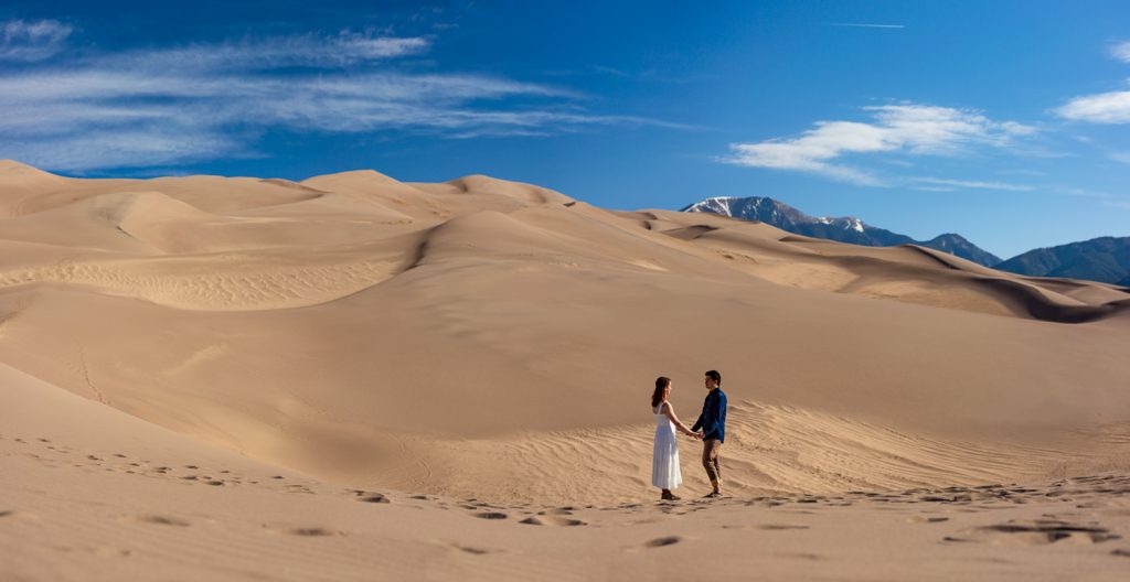 A man and a woman hold hands and look at each other while standing at the base of dramatic sand dunes under a bright blue sky.
