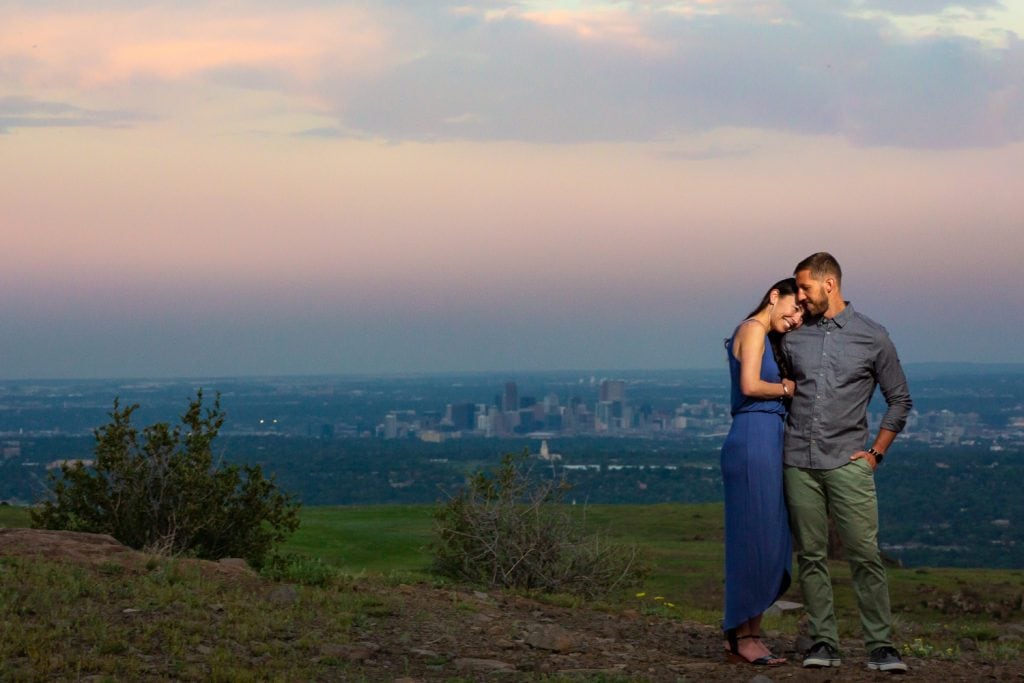 A couple cuddles up at sunset with the Denver city skyline in the background of their engagement photo.