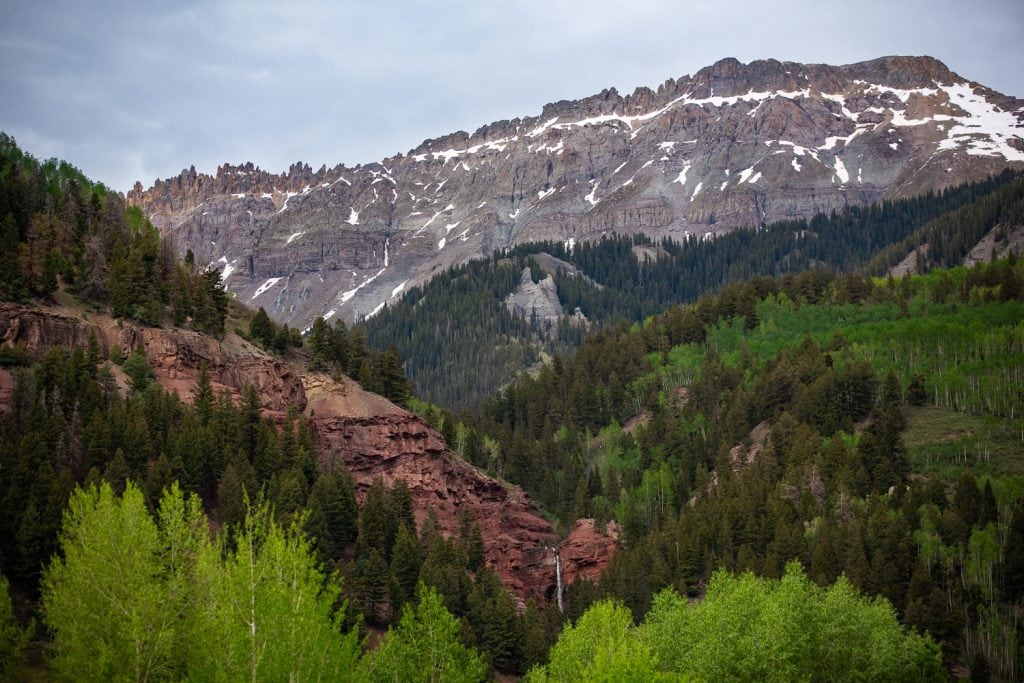 A waterfall, aspen trees, and exposed red rock form layers in front of Mt Sneffels near Telluride.