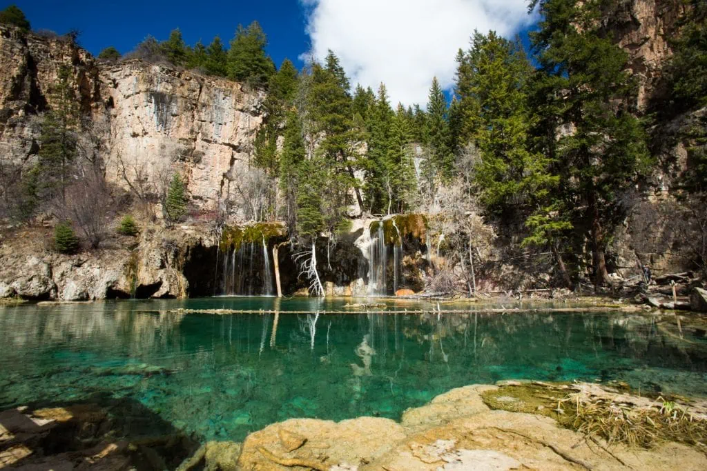 A colorful landscape photo of Hanging Lake, a unique geographic feature with a waterfall entering the turquoise water.