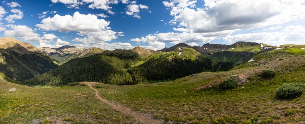 A huge panorama of mountain views in Central Colorado near Aspen. It it July and the terrain is green under a blue and cloudy sky.