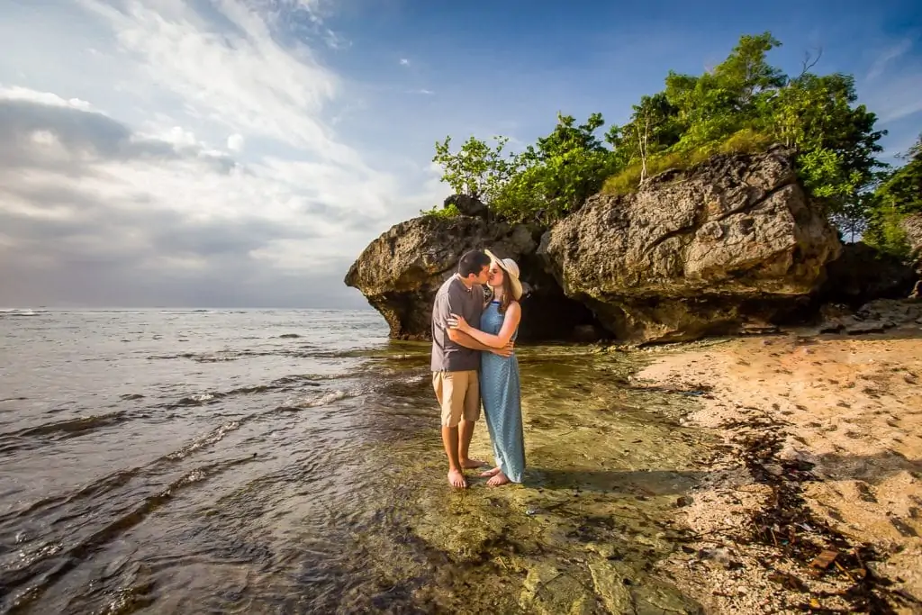 A couple kiss on the beach with tree-topped cliffs in the background by Bali photographer Lucy Schultz.