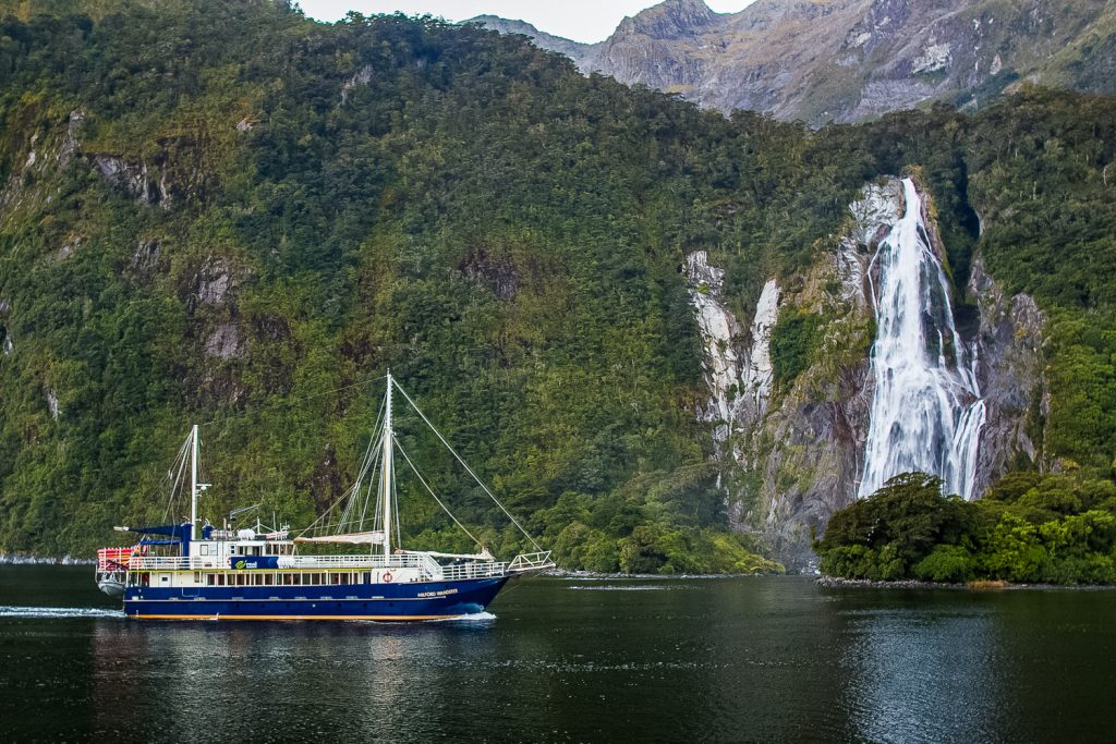 A sailboat cruise crosses Milford Sound in front of a waterfall.