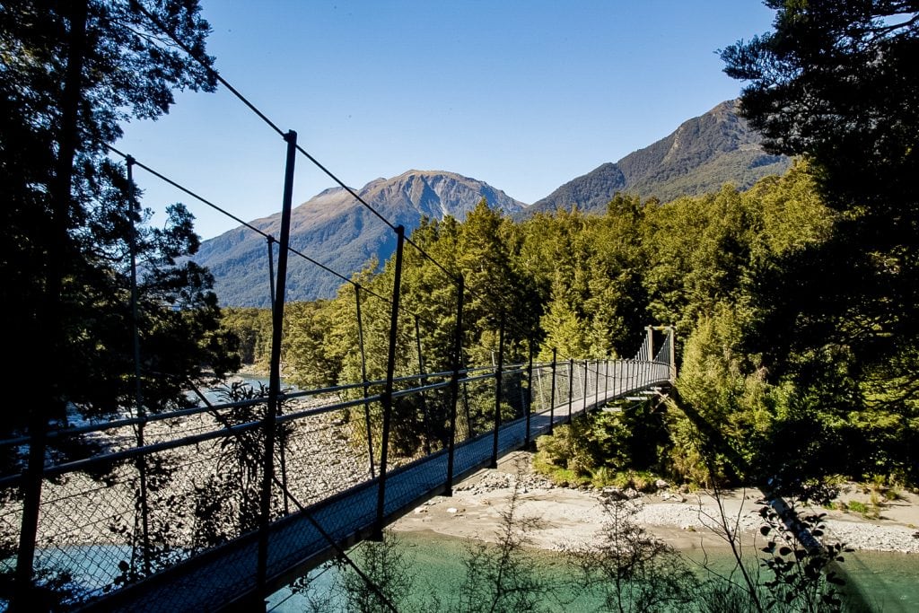 A hanging bridge over a river in New Zealand with mountains in the background.