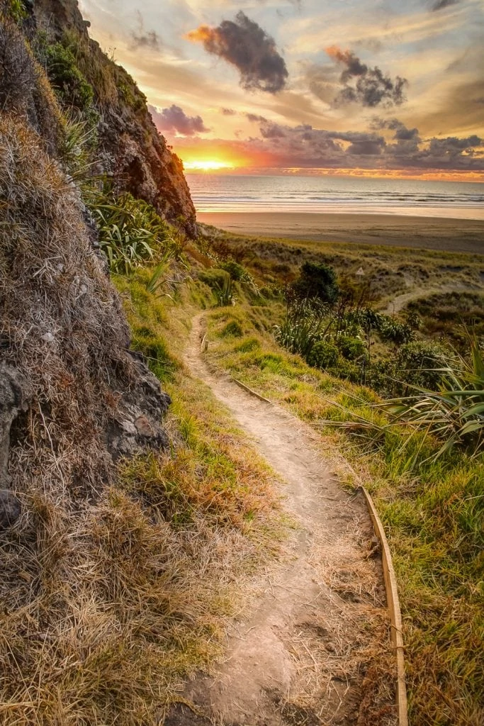 A coastal path alongside a mountain leads to an ocean beach at sunset.