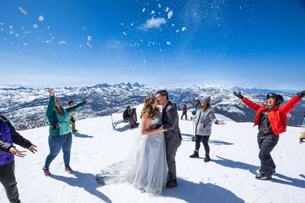 Bystanders throw snow over a n elopement couple at the top of a mountain.