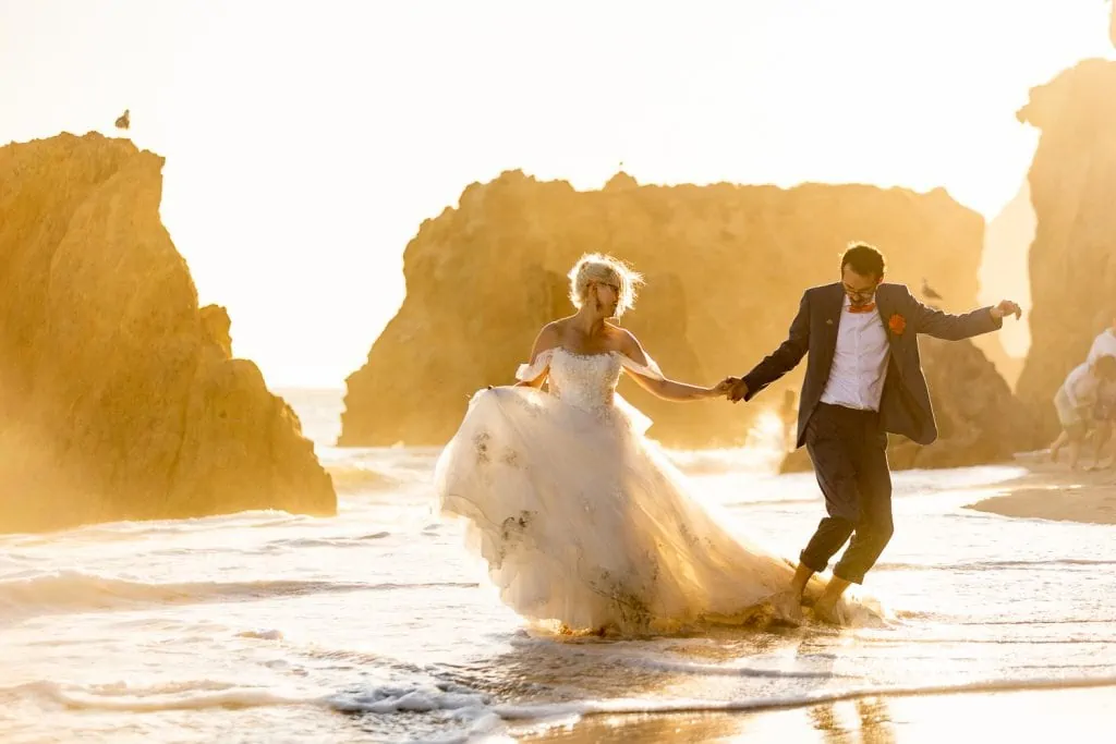 A wedding couple runs down the beach at sunset in Malibu, California.