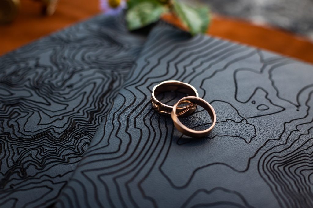 Two mens wedding rings sit on top of black vow books with topographic lines.