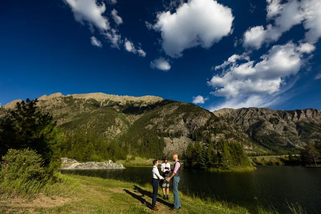 Two men hold hands as they exchange vows at a lake with a mountain in the background.
