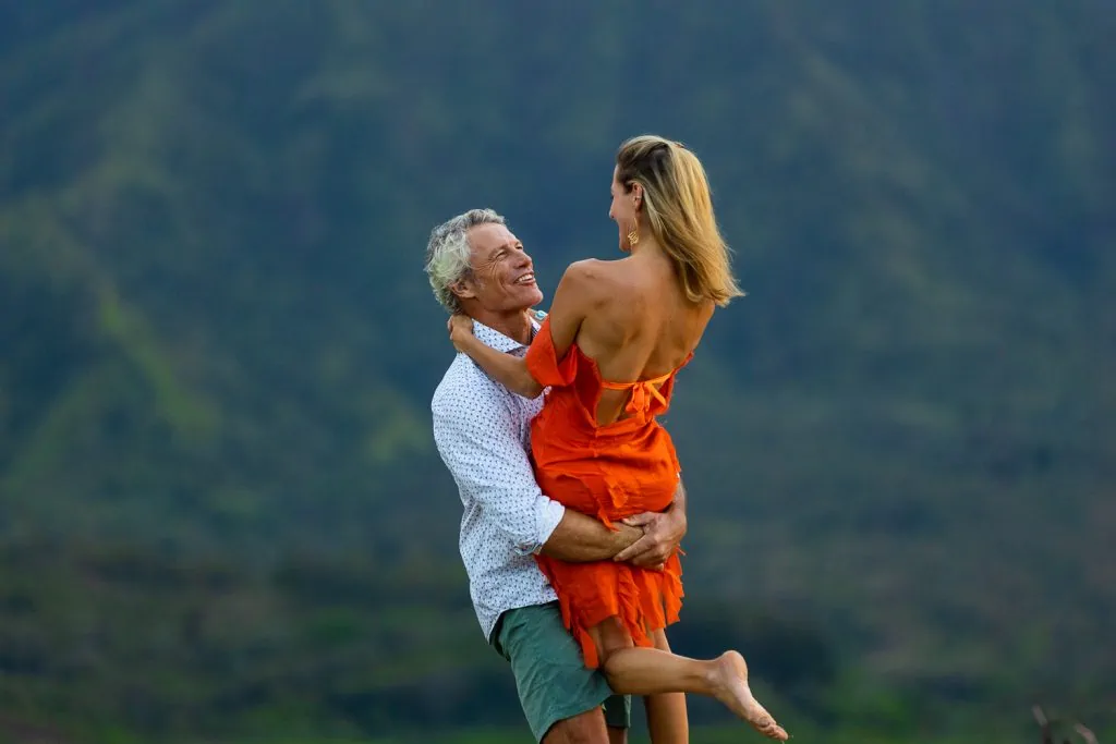 A handsome older man smiles at his wife as he lifts her and spins her around with the mountains of Kauai in the background.