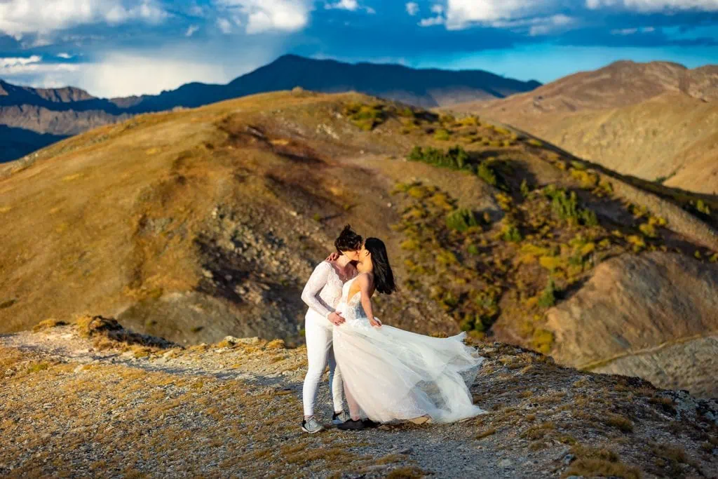 Two brides with windblown dresses share a kiss on top of a mountain in Colorado. The brides hiked for ten minutes to get to this vista.