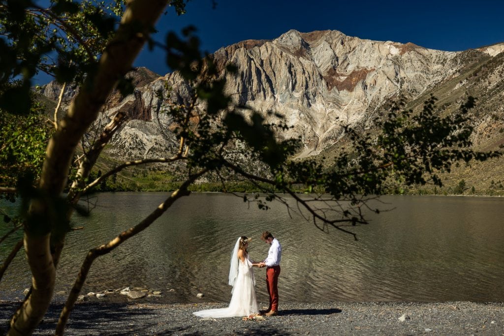 An elopement couple stands alone on the shore of a mountain lake in California for their just us two elopement photos.