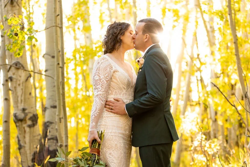 A bride and groom kiss in a grove of yellow aspens at sunset.