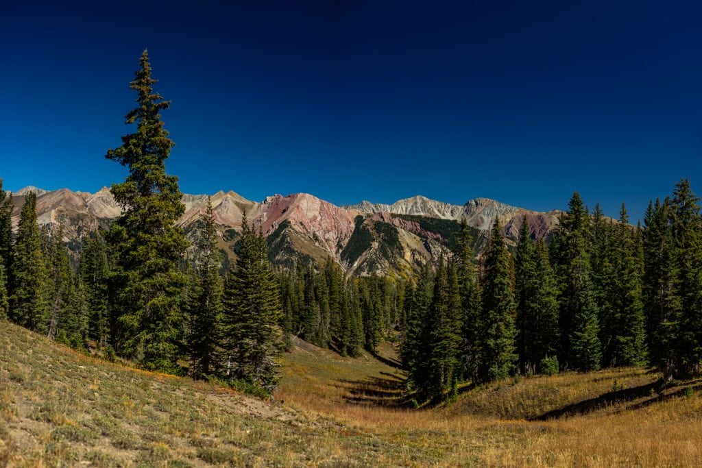 Red mountain peaks rise above evergreens in this landscape photo in Crested Butte, Colorado.