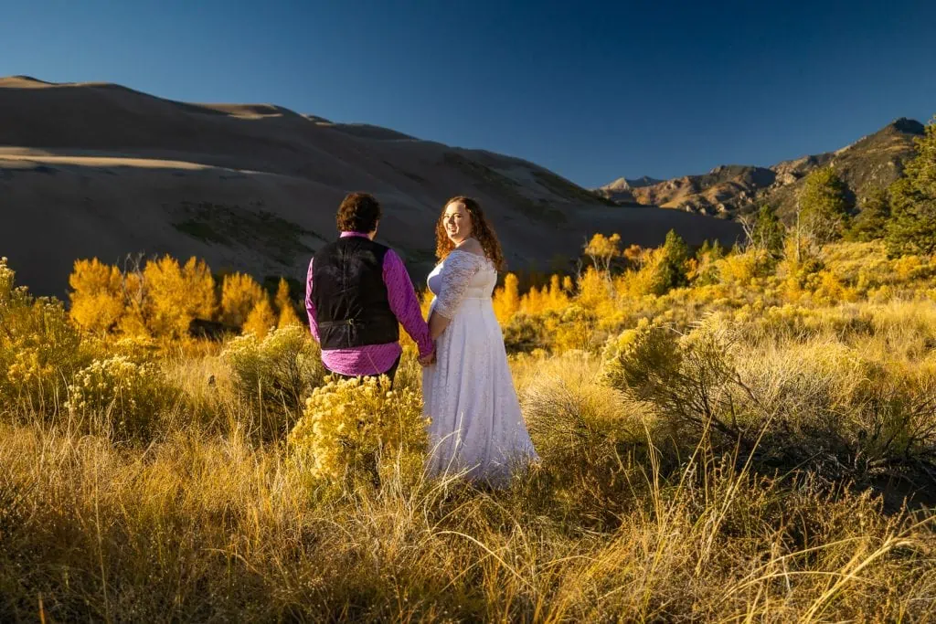 The bride looks over her shoulder at the camera while holding hands with her groom at their Great Sand dunes elopement.