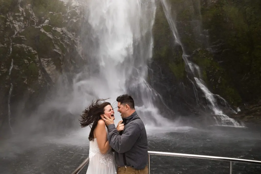 A candid moment of a bride and groom getting pummeled by a waterfall at their new zealand adventure elopement in fjordland national park. 