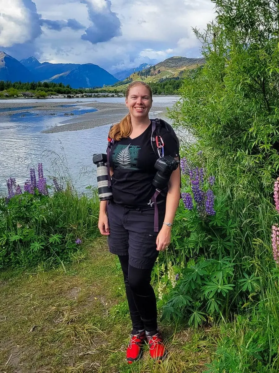 Worldwide elopement photographer Lucy Schultz is photographed here on assignment in New Zealand. She is wearing a backpack with 2 cameras, black clothes, and red shoes.