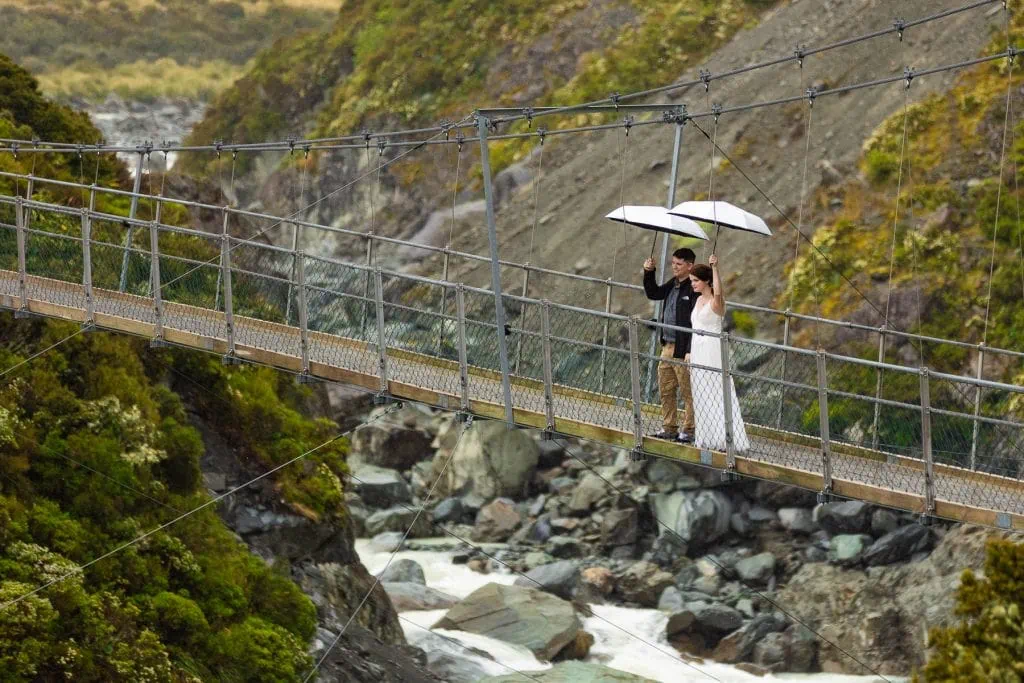 A wedding couple holding umbrellas on a swing bridge while hiking the hooker lake track in New Zealand. 