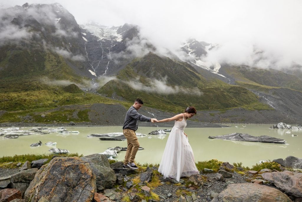 An adventurous elopement couple exploring the rocky shore of Hooker Lake with Mt Cook in the background in New Zealand.