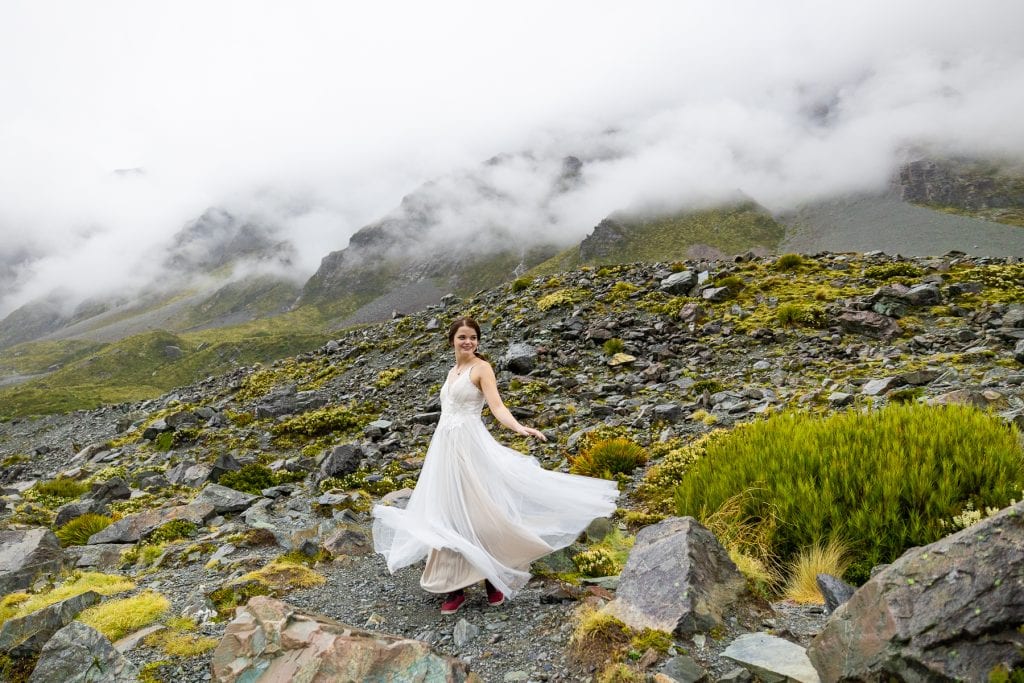 The bride twirls in front of a misty mountain range. Green and yellow alpine shrubbery are mixed in with dark rocks. 