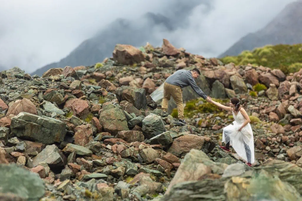 The groom helps a bride up a steep rocky slope. 