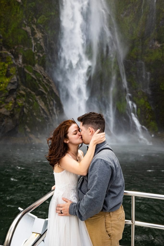 A passionate kiss between an adventure elopement couple with Stirling Falls in the background. The couple is standing on the front of a boat on the ocean.