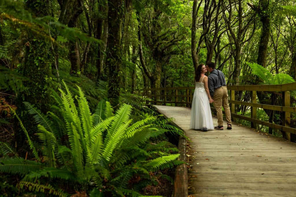 The groom kisses the bride's cheek in the fern jungle on a boardwalk on the south island of New Zealand. 