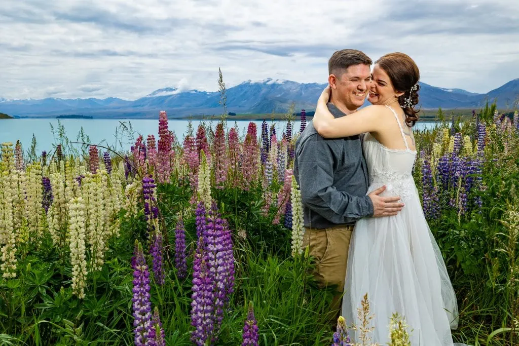 A wedding couple in purple and white lupine flowers on the shore of turquoise Lake Tekapo, New Zealand.