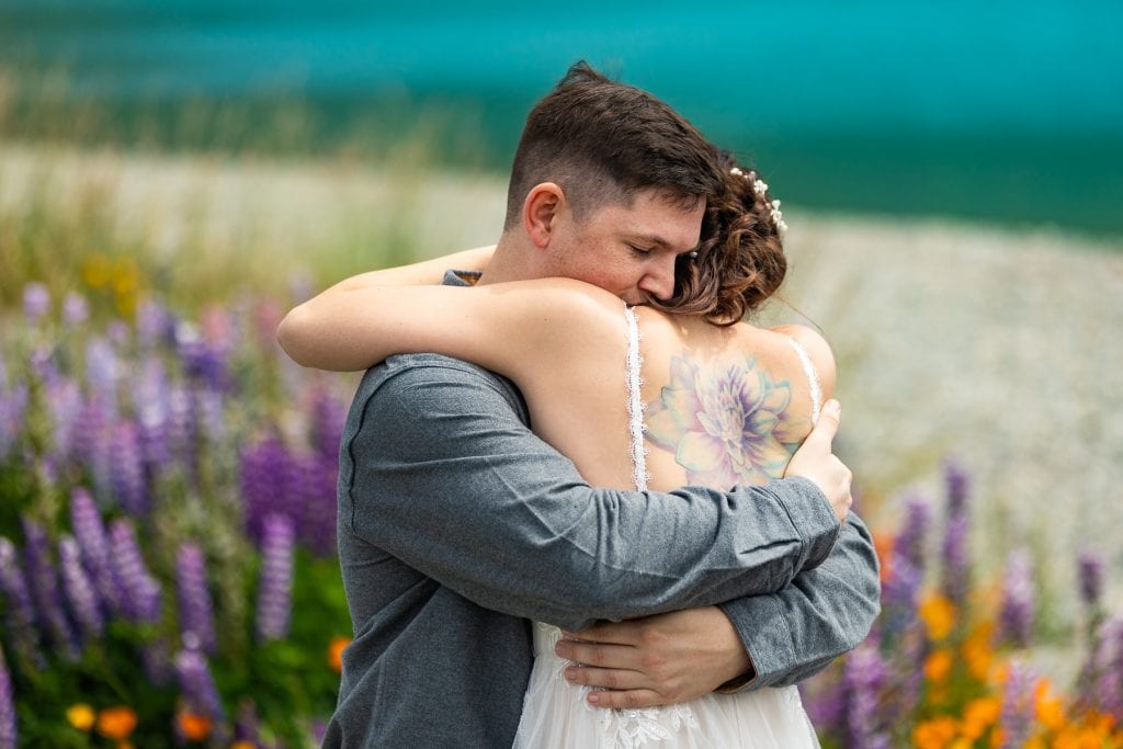 The groom embraces the bride while in a field of flowers in New Zealand during their elopement. Her watercolor tattoo on her back is framed by her dress.