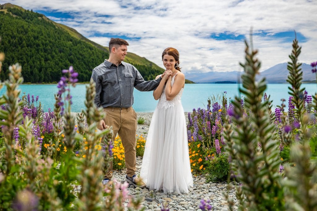 The gleeful bride clasps her new husband's hand in a field of flowers on the shore of Lake Tekapo, New Zealand. 