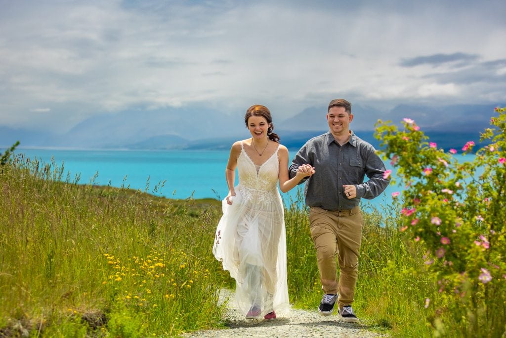 A wedding couple runs up a path with New Zealand's Lake Pukaki shining in the background. 