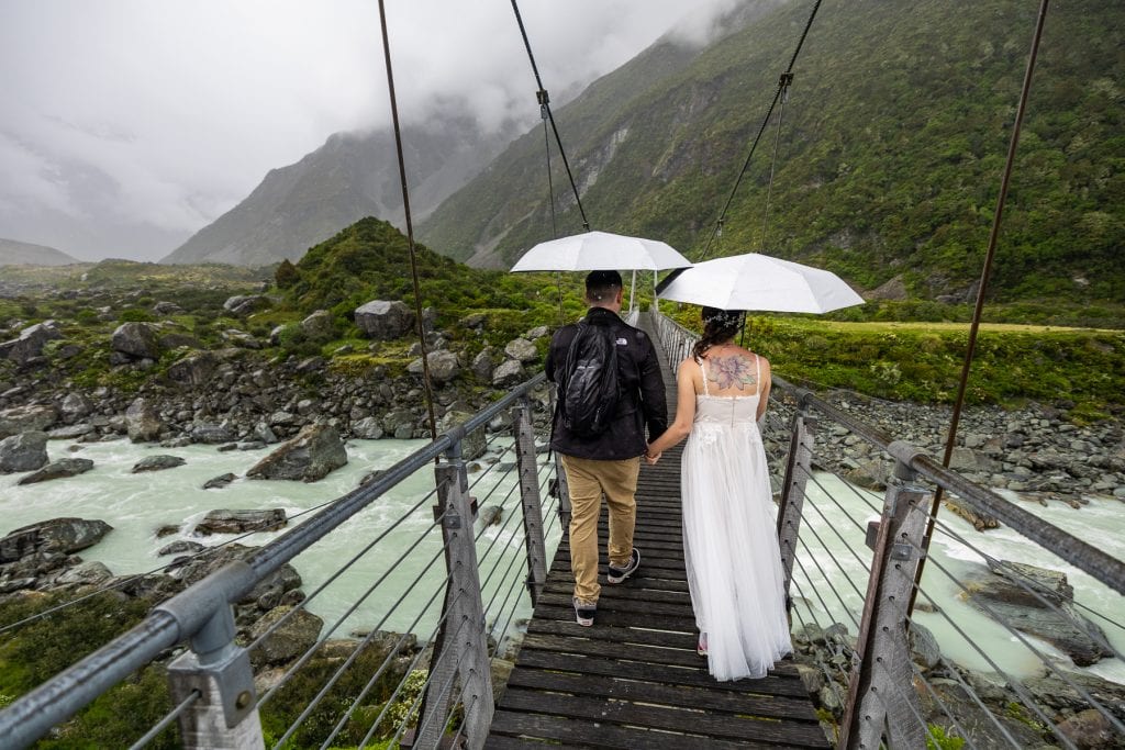 The wedding couple crosses a swing bridge while hiking in New Zealand on a rainy day. They are holding umbrellas. 
