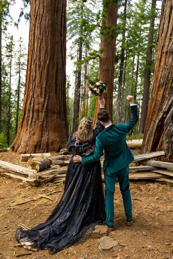 The ecstatic newlyweds pump their fists in the air after their yosemite elopement ceremony in the woods. The bride is wearing an edgy black leather jacket with Just Married printed on the back.