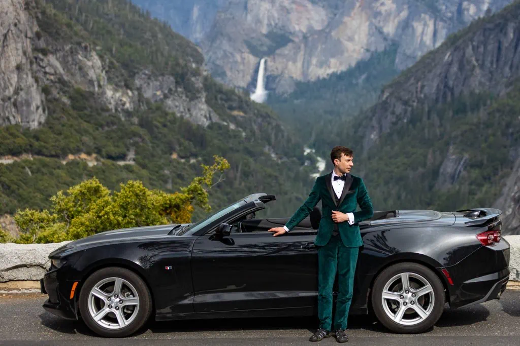 A dramatic photo of a groom wearing a green tuxedo posing in front of a black convertible with yosemite's waterfalls in the background.