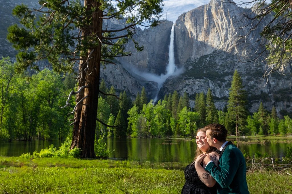 The groom snuggles up to his wife in front of yosemite falls, surrounded by green trees in this colorful Yosemite elopement photo.
